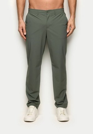 Mężczyzna w oliwkowozielonych spodniach i białych tenisówkach stoi na tle jednolitego tła, tułów częściowo widoczny.