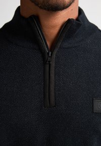 Maglione zip di colore nero con un design a maglia testurizzata e un colletto alto, dotato di una cerniera nera uniforme e di una piccola etichetta con logo di lato.