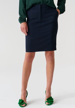 Woman wearing a dark navy knee-length pencil skirt, green blouse, and green open-toe heels with flower embellishments, standing against a plain background.