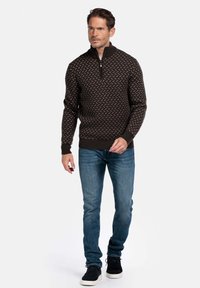 Man loopt in een geblokte, donkere ritssluitingtrui, blauwe jeans en zwarte sneakers met witte zolen op een witte achtergrond.