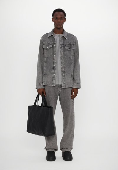 Veste en denim gris avec deux poches poitrine, associée à un pantalon gris assorti. L'individu tient un sac fourre-tout en cuir noir avec des poignées courtes.