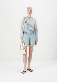 Pettorina in denim azzurro chiaro, con una sola spallina e orli arrotolati, abbinata a una felpa grigia, infradito nere e una borsa bianca.