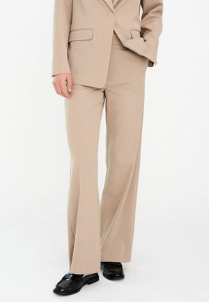 Bézs, testre szabott, széles szárú nadrág egyező blézerrel és fekete, fényes mokaszin cipővel, egy személy, akinek az egyik keze a zsebében van.