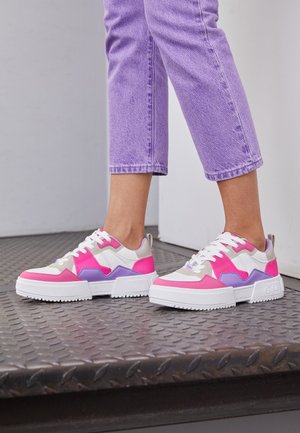 Par de pies con zapatillas blancas con detalles en rosa y púrpura, de pie sobre una superficie metálica texturizada, con vaqueros morados cortos.