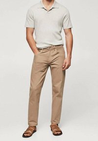 Homme portant un pantalon beige, un polo tricoté à manches courtes gris clair et des sandales en cuir marron, se tenant debout avec une main dans la poche.