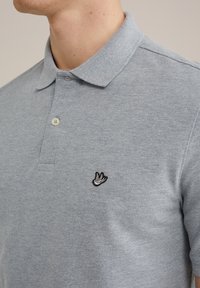 Pelēka polo krekls, kas izgatavots no teksturēta auduma, ar klasisku apkakli, divām pogām un izšūtu melnbaltuputnu logo uz krūtīm.