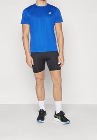 T-shirt de sport bleu à manches courtes, short noir ajusté, chaussettes blanches et chaussures de course noires avec des accents verts. Design simple et fonctionnel.