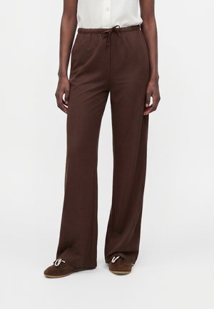 Femme portant un pantalon large marron à cordon et des chaussures marron, debout devant un fond clair uni.
