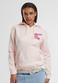 Helle rosa Kapuzenpullover mit vorderer Kängurutasche und Kordelzug. Mit dem Text "WARTEN AUF DEN FREITAG" in fettem Pink. Kombiniert mit einem silbernen Nieten-Gürtel.