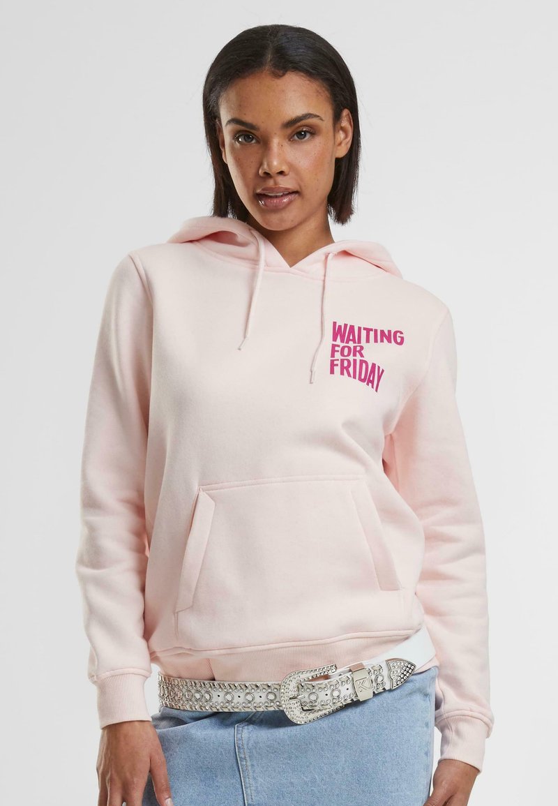 Helle rosa Kapuzenpullover mit vorderer Kängurutasche und Kordelzug. Mit dem Text "WARTEN AUF DEN FREITAG" in fettem Pink. Kombiniert mit einem silbernen Nieten-Gürtel.