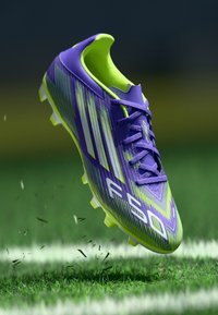 Crampons de football violet et vert lime avec un design texturé, comportant trois rayures blanches et un profil léger. Semelle extérieure à crampons.