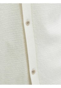 Εγγύς άποψη υφάσματος πλεκτού σε κρεμ απόχρωση με κάθετο κούμπωμα που διαθέτει δύο μπεζ κουμπιά.