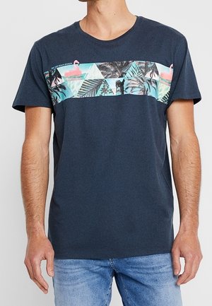 Uomo che indossa una maglietta blu navy con una striscia orizzontale a stampa tropicale e jeans azzurri, in piedi con le braccia rilassate lungo i fianchi.
