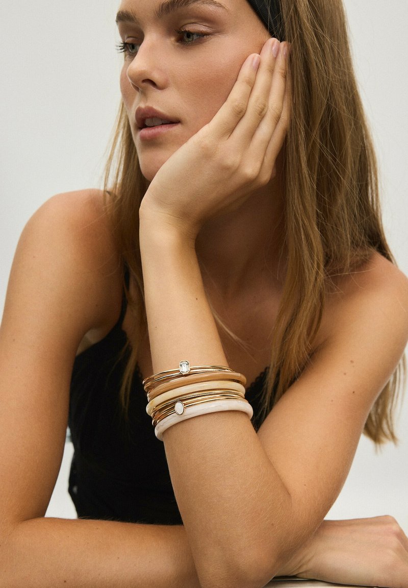 Femme reposant son visage sur sa main, portant des bracelets dorés et blancs superposés, avec de longs cheveux bruns et un haut noir sans manches.