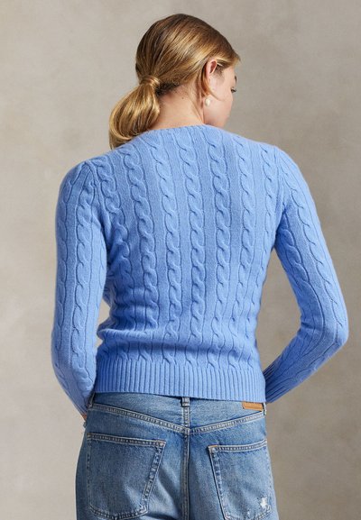 Woman with blonde hair in a ponytail wearing a light blue cable-knit sweater and blue denim jeans, facing away against a neutral background.