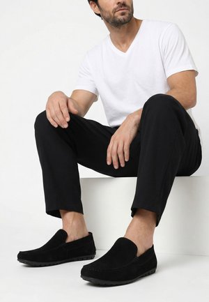 Mann sitzt auf einem weißen Block, trägt ein weißes V-Ausschnitt-T-Shirt, schwarze Hose und schwarze Slipper mit strukturierten Sohlen.