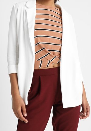 Kobieta ubrana w biały żakiet na beżowym i czarnym paskowanym krótkim topie, w połączeniu z wysokim stanem bordowymi spodniami.
