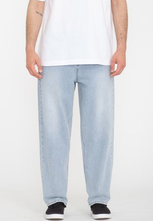 Light blue denim jeans feature a loose fit, straight legs, and minimal stitching. Paired with a plain white t-shirt and black sneakers.