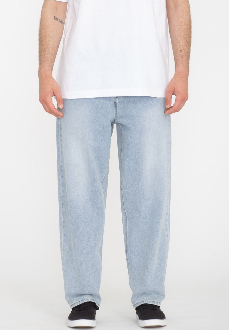 Light blue denim jeans feature a loose fit, straight legs, and minimal stitching. Paired with a plain white t-shirt and black sneakers.