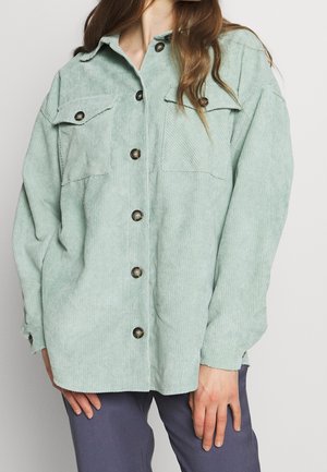 Femme portant une chemise en velours côtelé vert clair avec boutons et deux poches poitrine, ainsi qu'un pantalon violet foncé, debout devant un fond uni.