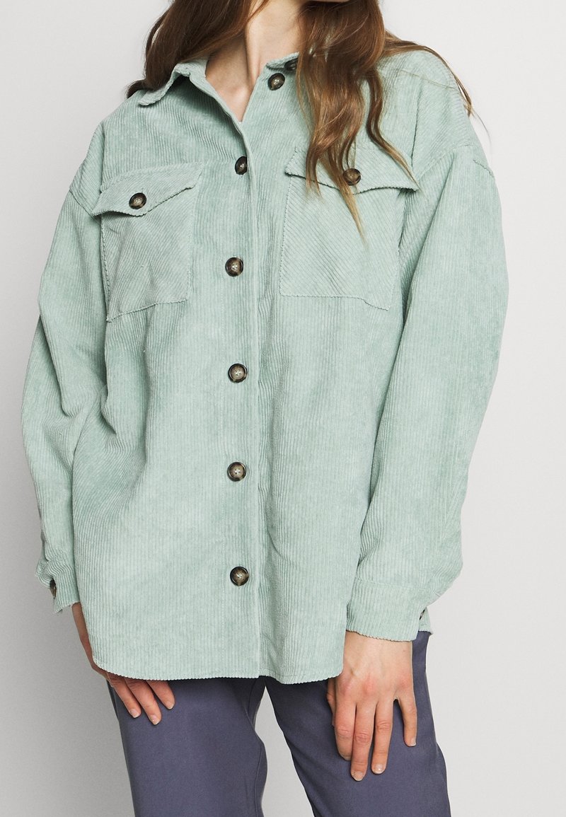 Femme portant une chemise en velours côtelé vert clair avec boutons et deux poches poitrine, ainsi qu'un pantalon violet foncé, debout devant un fond uni.