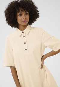 Svetlo béžové šaty s golierom, krátkymi rukávmi, zapínaním na gombíky s hnedými gombíkmi a voľným strihom pre pohodlie.