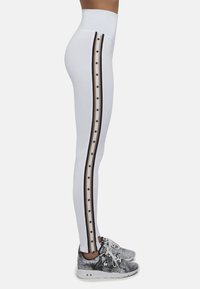 Białe legginsy z wysokim stanem, z bocznymi pasami w kolorze czarnym i beżowym oraz wzorami gwiazdek; zestawione z fakturowanymi szarymi sneakersami.