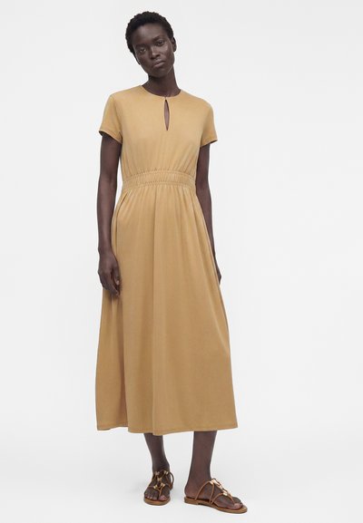 Femme aux cheveux courts portant une robe midi beige à manches courtes avec un col keyhole et des sandales plates marron, debout devant un fond uni.