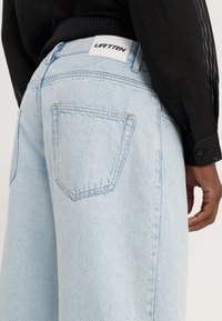 Jean en denim bleu clair avec une coupe décontractée, doté d'une étiquette en cuir blanc marquée "UR TRN" et de deux poches arrière avec une finition légèrement texturée.