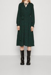 Mörkgrön knälång klänning med litet turkost blommönster, v-ringning med knytband, långa ärmar och skärp i midjan. Bärs med svarta knähöga stövlar.