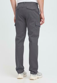 Man wearing dark gray cargo pants with back and side flap pockets, white sneakers, standing against a plain light background.