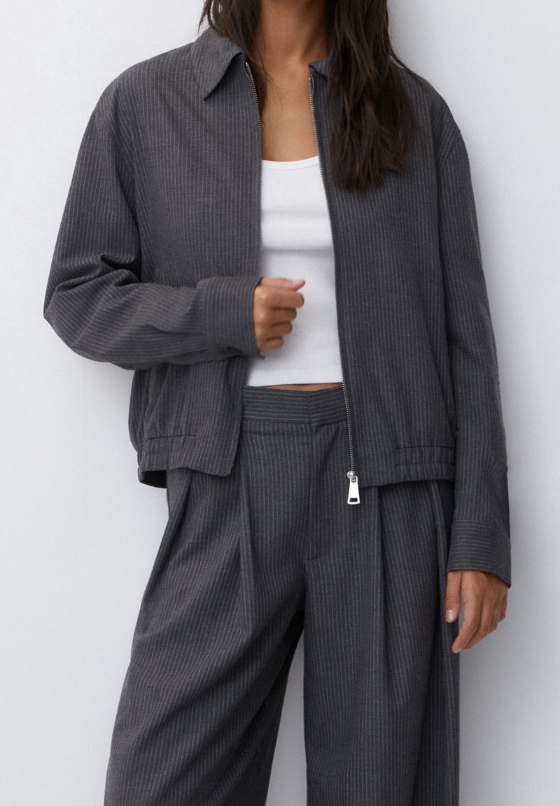 Femme portant une veste zippée gris foncé à fines rayures et un pantalon large assorti sur un top court blanc.