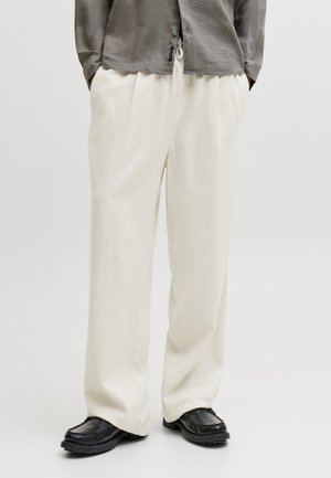 Mand iført løsthængende off-white bukser, sorte læderloafers og en grå langærmet skjorte med hænderne i lommerne.