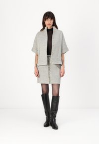 Giacca corta grigia in tessuto tweed con taglio dritto, due grandi bottoni, abbinata a una gonna alta in vita dello stesso materiale e stivali neri fino al ginocchio.