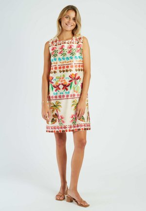 Woman standing and smiling, wearing a sleeveless dress with colorful floral and palm tree patterns, and beige heeled sandals.