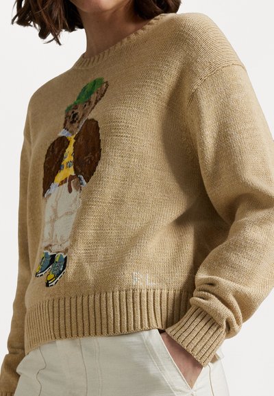 Beige gebreide trui met een beerafbeelding met een groene hoed, gele shirt en sneakers; geribbelde zoom en manchetten, zachte textuur.