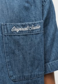 Τσέπη πουκάμισου από τζιν μπλε ύφασμα με λευκό κεντημένο κείμενο "Originals Studio" σε ένδυμα με κοντά μανίκια.