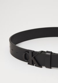 Cintura in pelle nera con una texture liscia, dotata di una notevole fibbia nera in rilievo con la scritta "CK" e un design elegante.