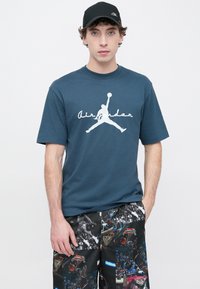 Μπλε σκούρο βαμβακερό t-shirt με λευκό λογότυπο Air Jordan. Συνοδεύεται από μαύρο σορτς με πολύχρωμο μοτίβο γραφικών μπάσκετ.