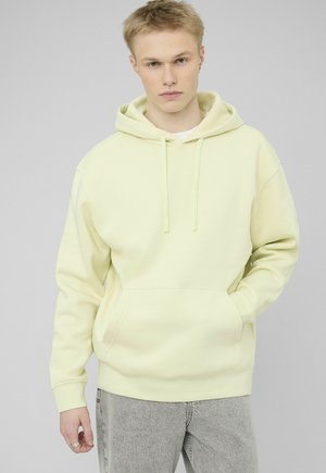 Jeune homme portant un sweat à capuche jaune pâle avec poche avant et un jean gris clair, debout devant un fond uni.