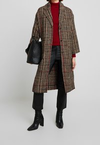 Lungo cappotto scozzese nei toni di marrone, nero e rosso, con colletto a picco, bottoni doppi, abbinato a pantaloni in pelle nera e stivali con tacco.