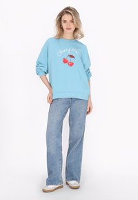 Ljusblå sweatshirt med körsbärsgrafik och texten "Cherry Me", i kombination med ljusblå vida jeans och vita sneakers.
