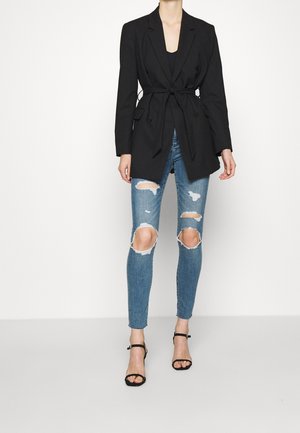 Black tailored blazer with a tie waist, double-breasted design, worn over light blue ripped skinny jeans and black strappy heels.