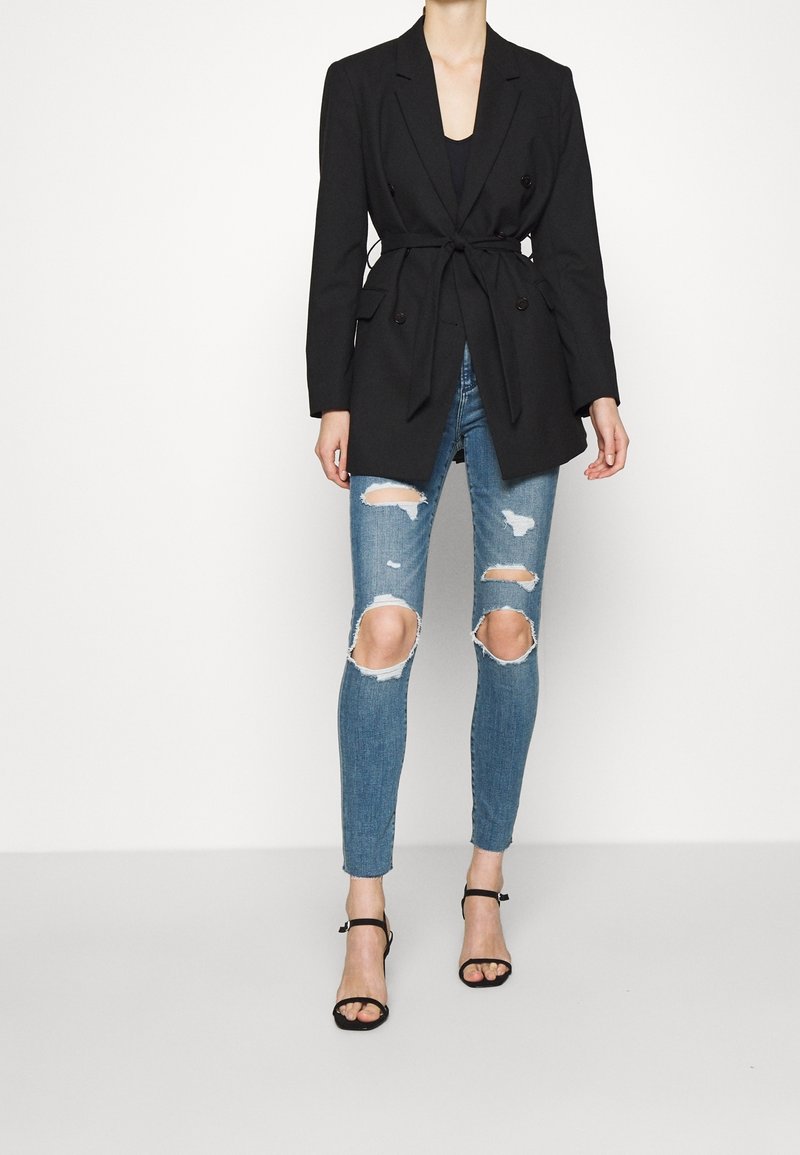 Black tailored blazer with a tie waist, double-breasted design, worn over light blue ripped skinny jeans and black strappy heels.