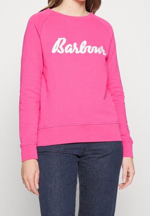 Personne portant un sweat-shirt rose vif avec un texte blanc "Barbour" et un jean bleu foncé, présenté du cou aux hanches sur un fond uni.