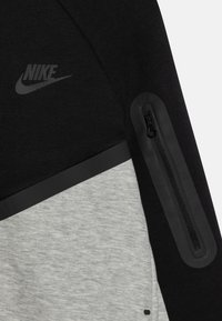Crni i sivi flis dukserica s velikim Nike logotipom, džepom sa zatvaračem na ruci i kontrastnim panele od tkanine. Teksturirana završna obrada.