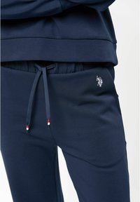 Tumši zili sporta bikses ar elastīgu jostu un auklu, kas piedāvā sarkanus, baltus un melnus akcentus; izšūts logo sānos.