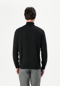 Schwarzer Rollkragenpullover aus glattem Strickmaterial; mit langen Ärmeln, figurbetontem Design und gerippten Bündchen sowie Saum. Getragen mit grauen Hosen.