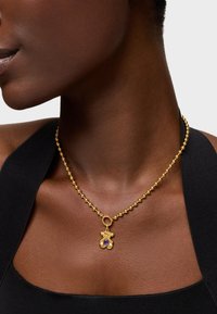 Collana di perline d'oro con un pendente centrale che presenta una pietra preziosa viola, appesa a un colletto liscio e scuro.