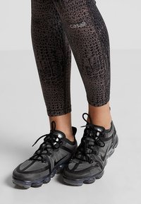 Czarno-szare legginsy sportowe z wzorem krokodyla i logo z boku, w połączeniu z czarnymi trampkami Nike z przezroczystą podeszwą.